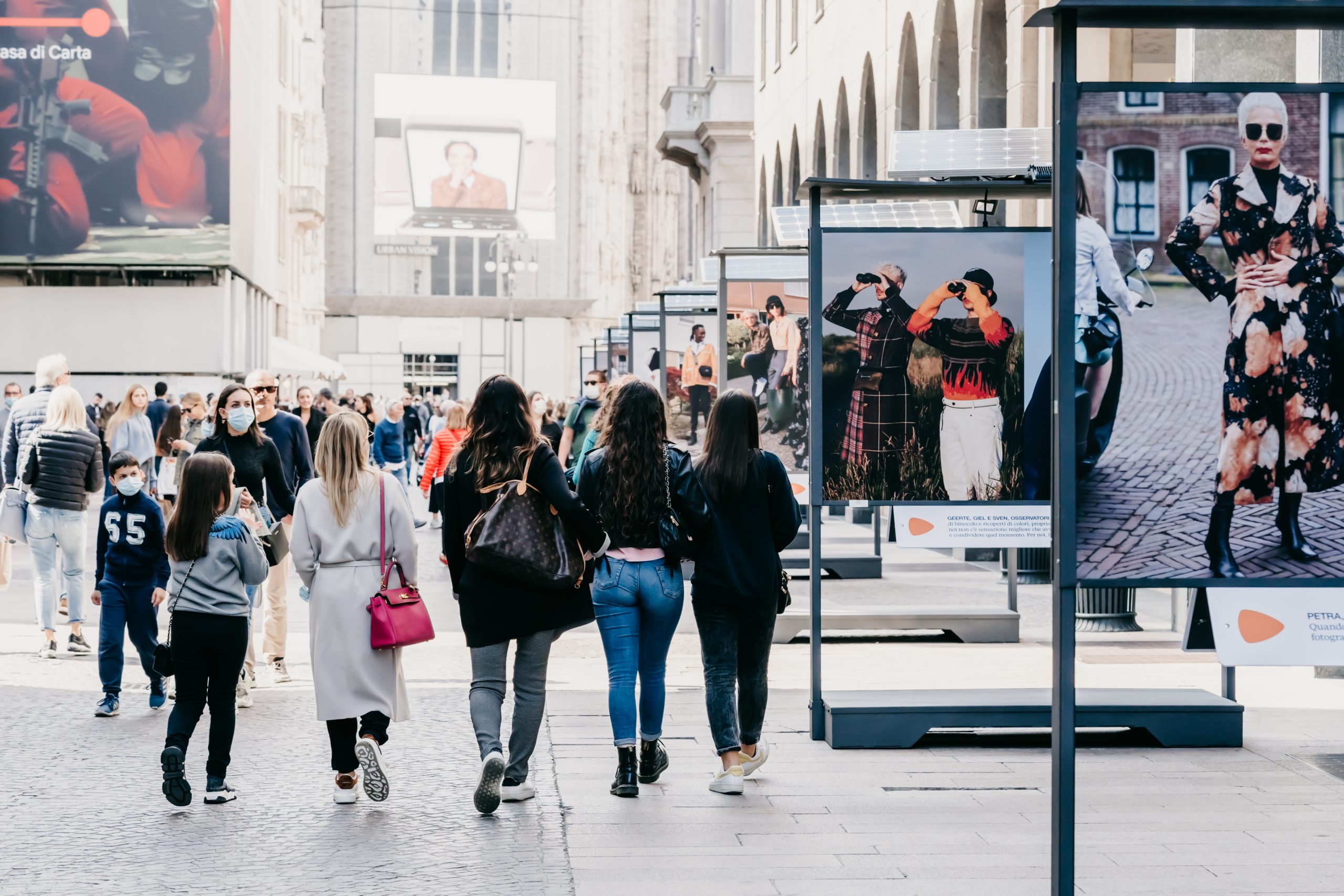 People in masks walking street in downtown of Milan, commuting, looking at modern fashion photography street exhibition using solar batteries. New normal covid